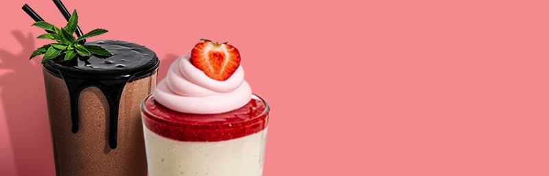 Two tall Keto Chow shakes in front of a coral-pink background—one chocolate shake topped with chocolate syrup, mint leaves, and black straws; the other a vanilla shake with strawberry puree, swirled pink whipped topping, and half a strawberry.