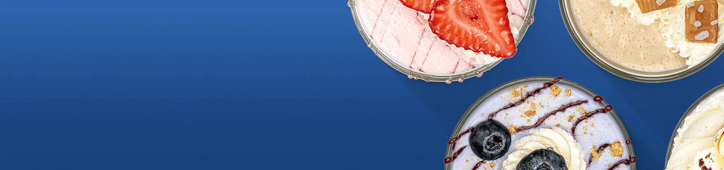 Four circular dessert bowls with various toppings are on a solid blue background. The top left bowl has pink contents with strawberries. The top right bowl has tan contents with cookies and whipped cream. The bottom left bowl has light purple contents with blueberries, whipped cream, and chocolate syrup. The bottom right bowl has yellow contents with whipped cream.