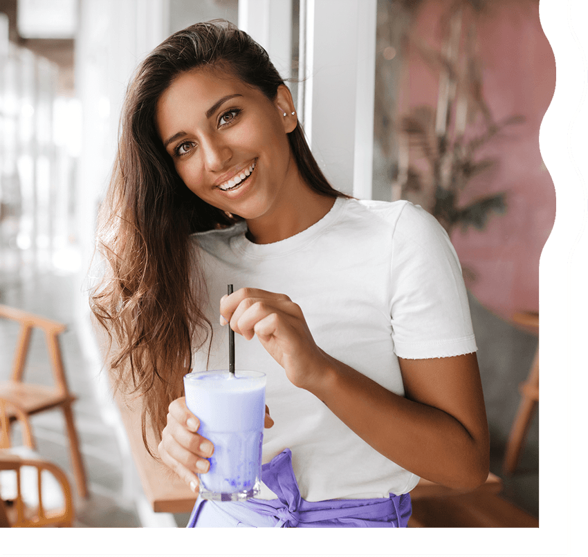 A woman with tan skin and long, dark brown hair is smiling and looking directly at the camera. She is holding a creamy, light blueish purple shake.