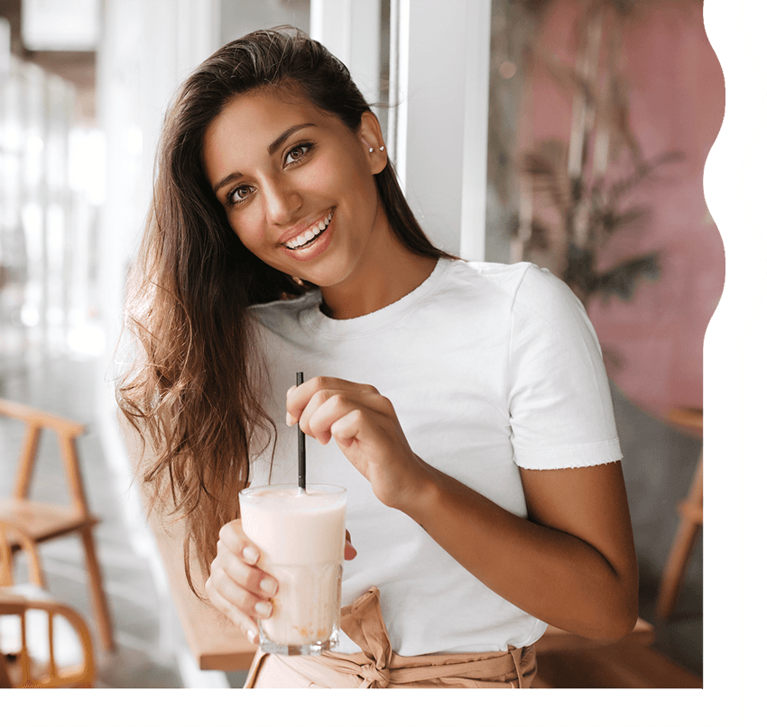 A woman with tan skin and long, dark brown hair is smiling and looking directly at the camera. She is holding a white, creamy shake.