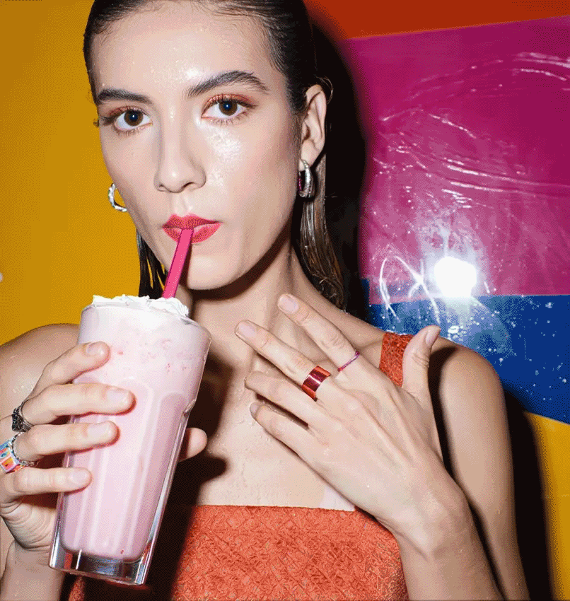 A woman with tan skin and long, dark brown hair is smiling and looking directly at the camera. She is holding a delicately pinkish red shake.